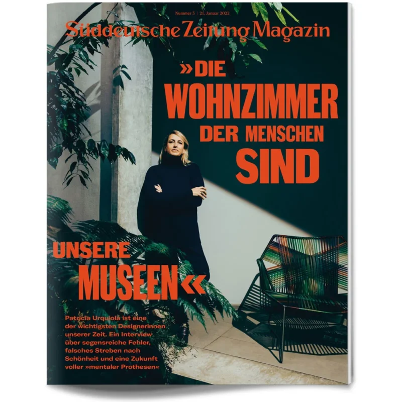 Eine schwarz gekleidete Frau steht in einem Innenraum zwischen Pflanzen; ein fetter orangefarbener deutscher Text bedeckt die Titelseite der Zeitschrift.