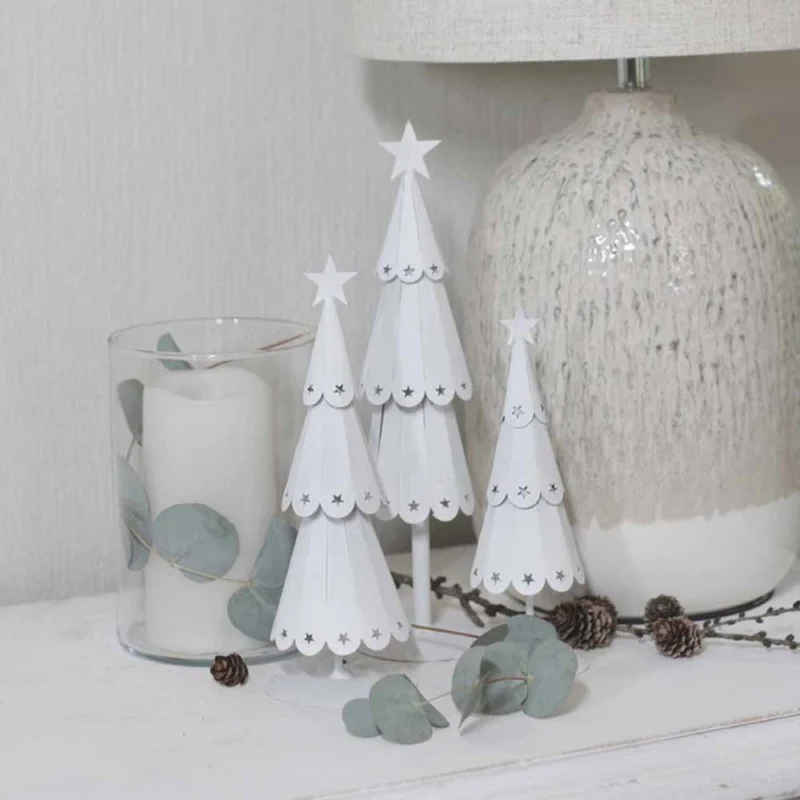 Der VL Home – Tannenbaum Metall mit Sternen und ein mit Eukalyptusblättern geschmückter Kerzenhalter aus Glas sind auf einer weißen Oberfläche neben einer strukturierten Lampe arrangiert.